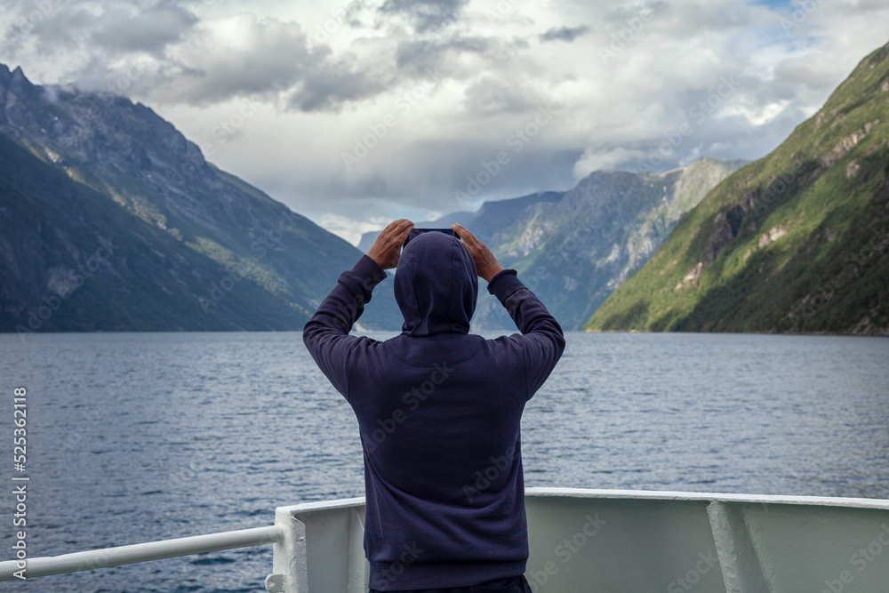 Norwegenreise - Tourist steht am Heck einer Fähre und macht mit dem Handy Fotos der atemberaubenden Bergwelt mit dem Geirangerfjord Norwegenreise - Tourist steht am Heck einer Fähre und macht mit dem Handy Fotos der atemberaubenden Bergwelt mit dem Geirangerfjord