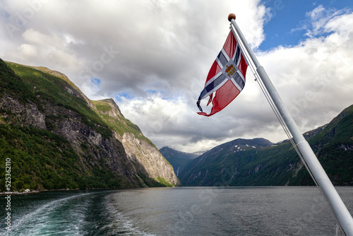 Norwegenreise - norwegische Flagge weht am Heck eines Schiffes mit Blick auf den Geirangerfjord und die atemberaubende Bergwelt -