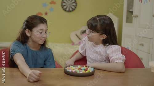 Little girls take a break from school homework classes and eat colorful candies. Little girlfriends eating colorful candies on the table in her room, laughing and having fun together.