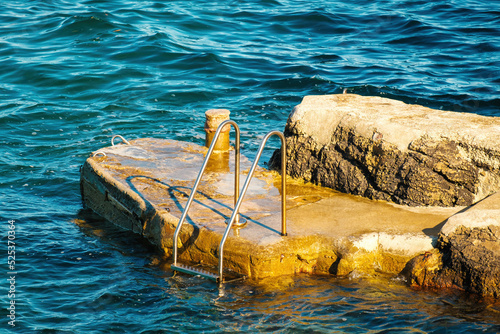 Fotografering Metal ladder for swimmers descending into sea from small rock at sunlight