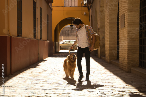 Tableau sur toile Young Hispanic man with beard and sunglasses with his dog holding on to the leash walking along a sunny street at sunset