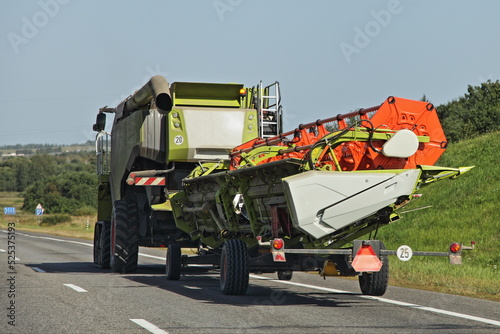 Modern European Harvester combine drive with reaping machine on trailer on countryside asphalt road at summer day - harvesting, agriculture, farming in East Europe
