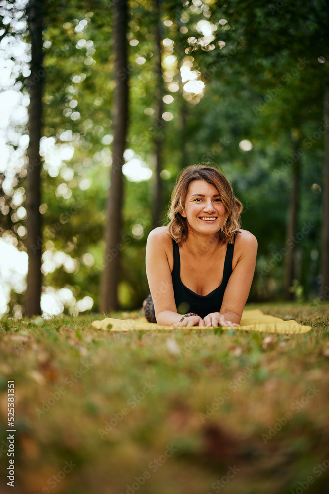 A happy woman is lying on her stomach on a yoga mat in the forest and taking a break from exercising. A happy woman is lying on her stomach on a yoga mat in the forest and taking a break from exercising.