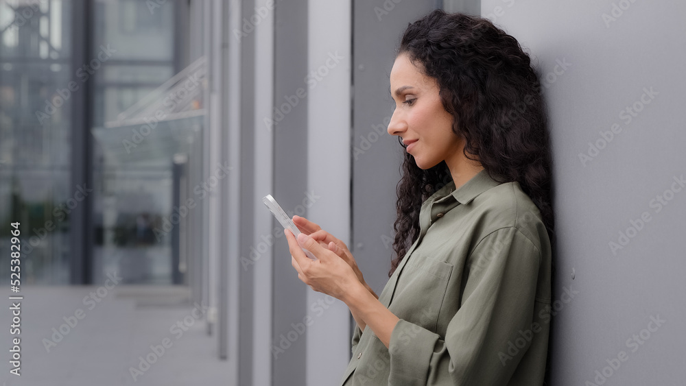 Fototapeta premium Latin Caucasian Hispanic girl woman 30s businesswoman holding cell phone standing near wall building outdoors success good news mobile app win surprised lady celebrating victory on smartphone triumph