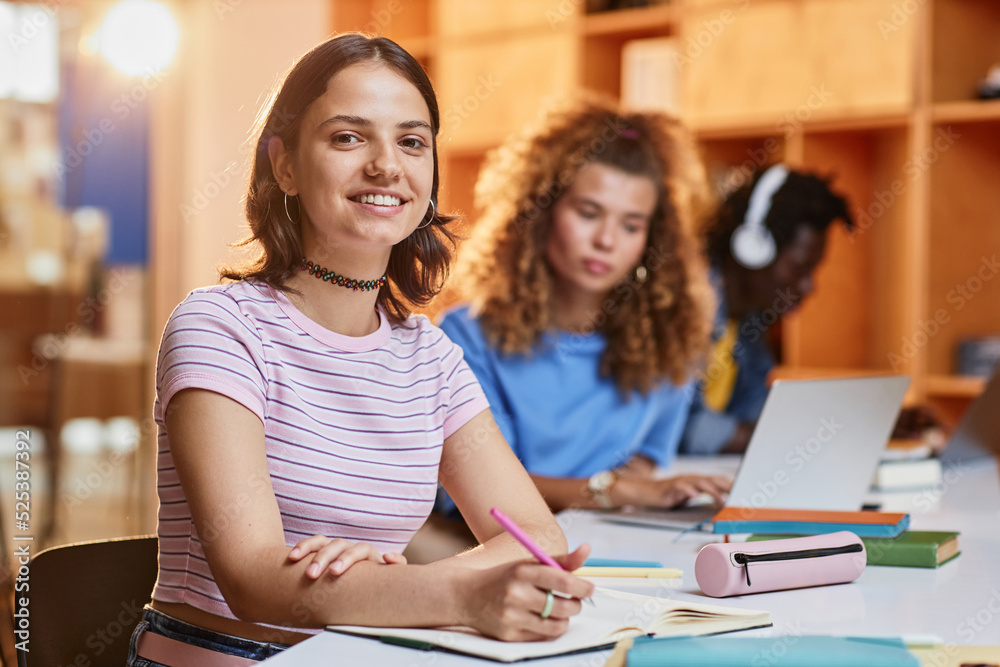 Diverse group of students in row studying in college library, focus on smiling young woman looking at camera in foreground, copy space Diverse group of students in row studying in college library, focus on smiling young woman looking at camera in foreground, copy space