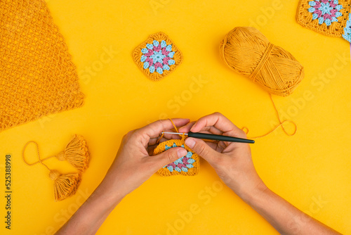 Top view of hands crocheting square element on yellow background with different crocht patterns, ball of yarn and yellow tassels around.