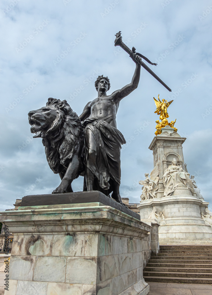 London, England, UK - July 6, 2022: Victoria Memorial. Closeup of Black bronze Progress statue of lion, youth with flaming torch under blue cloudscape. Central marble monument in back