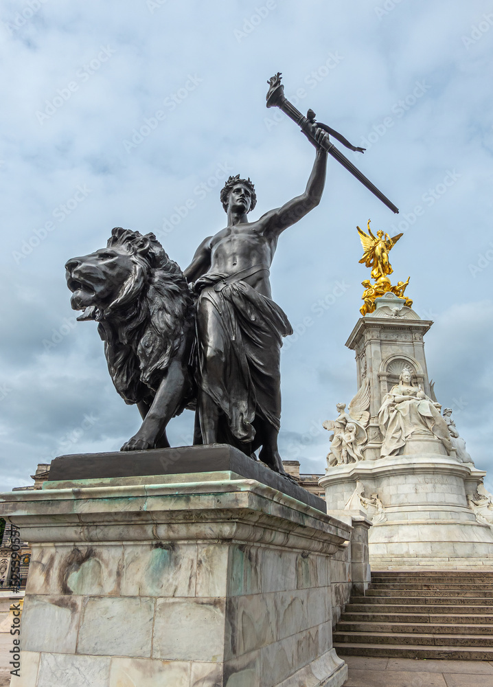 Obraz premium London, England, UK - July 6, 2022: Victoria Memorial. Closeup of Black bronze Progress statue of lion, youth with flaming torch under blue cloudscape. Central marble monument in back