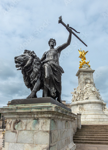 London, England, UK - July 6, 2022: Victoria Memorial. Closeup of Black bronze Progress statue of lion, youth with flaming torch under blue cloudscape. Central marble monument in back
