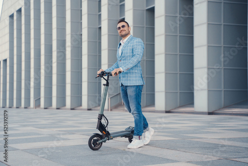 Joyful businessman with scooter dressed in blue suit and sunglasses against city building. © Fxquadro