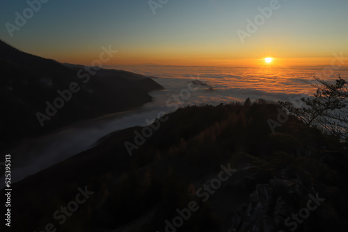 Sun rising above low clouds in mountain with dark hills in background. Nature scenery early in the morning with illuminated sky and valley still hidden in shadow.
