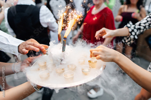 A tray of alcoholic drinks decorated with smoke and candles at a party. An outdoor party and a beautiful serving of alcohol for guests with liquid nitrogen in the form of smoke and sparks from candles photo