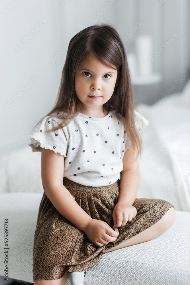 A cute little girl is sitting on a bed in a bright room. A serious child is dressed in a skirt and a T-shirt. Beautiful portrait of a little girl in her bedroom A cute little girl is sitting on a bed in a bright room. A serious child is dressed in a skirt and a T-shirt. Beautiful portrait of a little girl in her bedroom