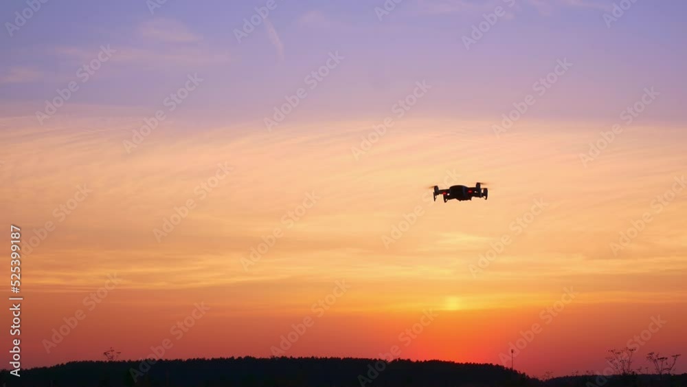 The drone moves from side to side in the bright sunset sky. The silhouette of a moving drone in flight with blinking lights against an orange sky flooded with sunset light. Concept of new technologies The drone moves from side to side in the bright sunset sky. The silhouette of a moving drone in flight with blinking lights against an orange sky flooded with sunset light. Concept of new technologies