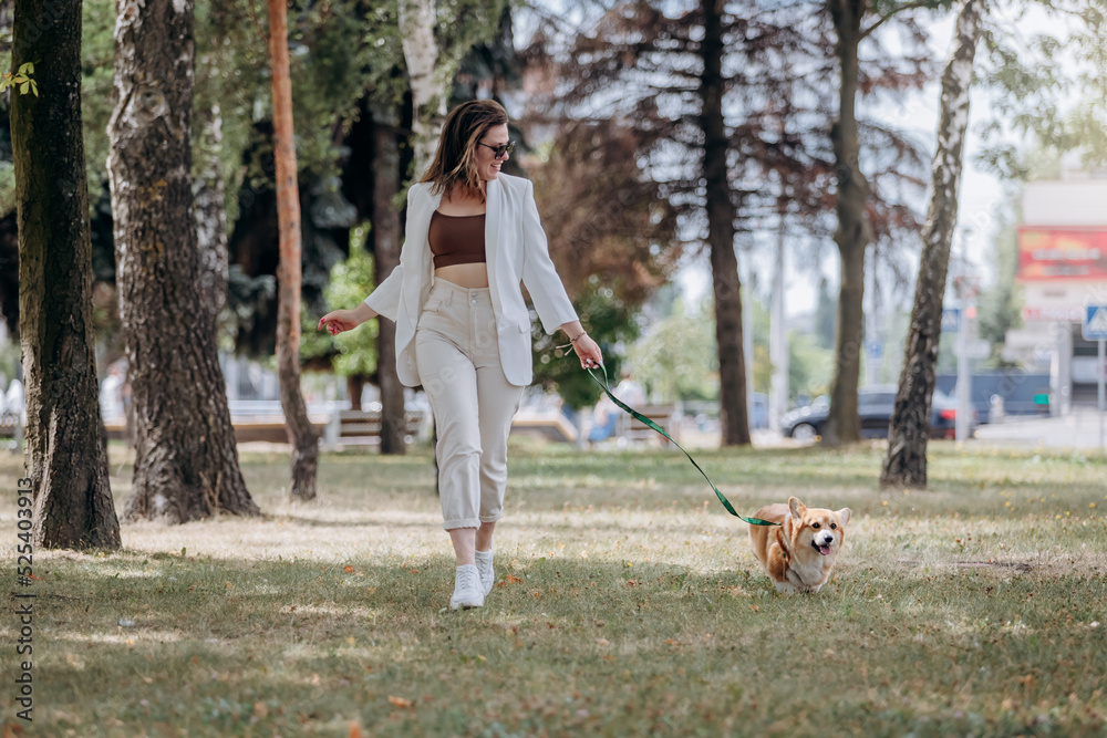 Happy Woman wearing white suit and sunglasses walking in city parkland with Welsh Corgi Pembroke dog