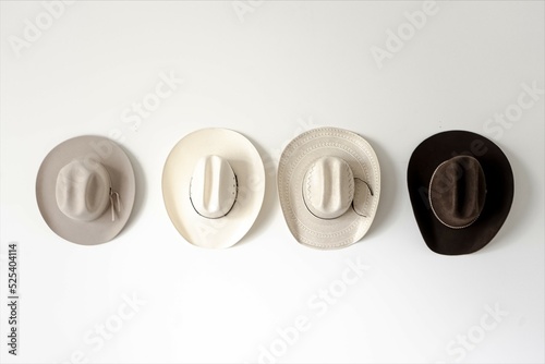 Four cowboy hats hung on a wall in various shades of white and brown against a white background with natural lighting.