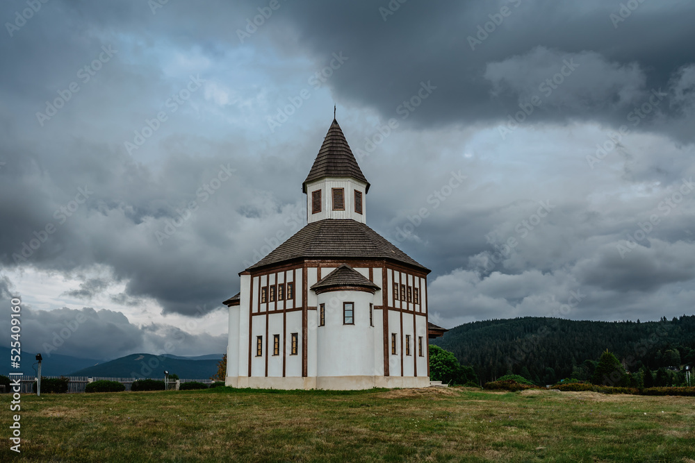 Fototapeta premium Small white rural Tesarovska chapel with cemetery in village of Korenov, Jizera mountains, Czech Republic. Summer landscape, stormy sky.Wooden historic church. Religious scenery.Czech countryside