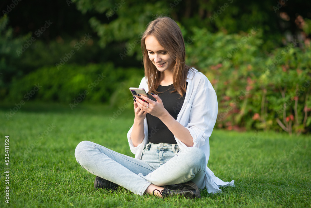 A young and attractive Caucasian girl in casual clothes is sitting on the grass in the park and using a mobile phone. A young and attractive Caucasian girl in casual clothes is sitting on the grass in the park and using a mobile phone.