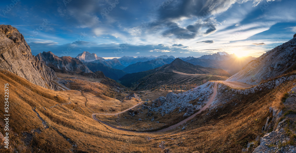 Beautiful view on mountains at sunset in autumn. Nature in Dolomites, Italy. Colorful panoramic landscape with rocks, orange grass in hills, trail, dirt road, stones, blue sky with clouds in fall