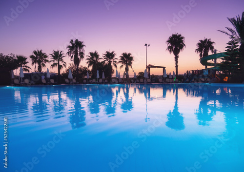 Beautiful reflection in swimming pool at colorful sunset. Purple sky reflected in water, palm trees, sun beds, umbrellas at night in summer. Luxury resort. Landscape with empty pool in twilight © den-belitsky