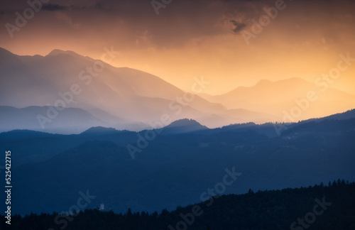 Amazing silhouettes of a mountains at colorful sunset in summer in Slovenia. Landscape with mountain ridges in fog, golden sunlight and clouds in the evening. Nature. Hills in sunlight. Scenery