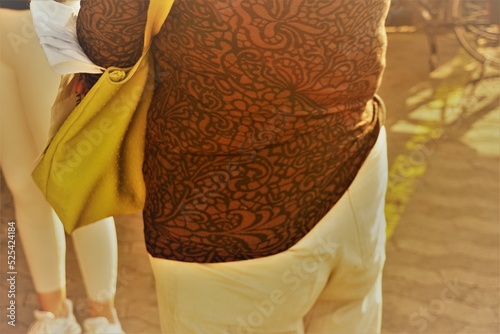 Rücken von Frau mit brauner Bluse mit Muster, beiger Hose und gelber Ledertasche mit weißer Corona-Maske in Stadt bei Sonne am Abend im Sommer