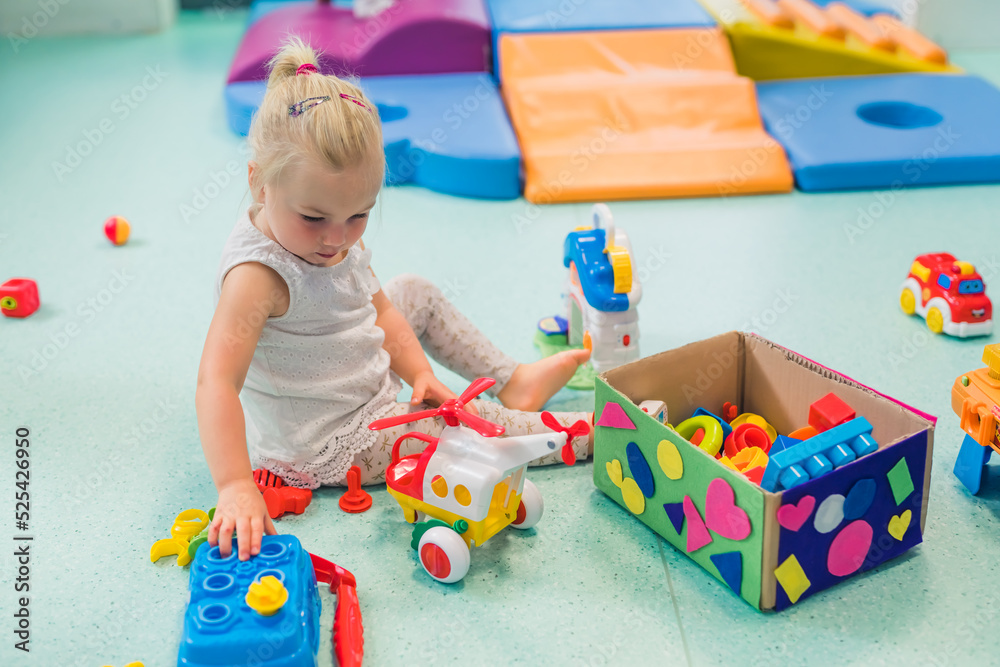 Fototapeta premium Toddler girl playing with different plastic toys such as building blocks, car toys. Work on problem-solving skills, shape and color recognition, fine motor and gross motor skills development through