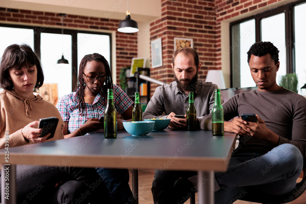Multiracial friends sitting at table in living room using on smartphones while surfing internet. Diverse group of people sitting at home while using modern mobile phones to watch video content.