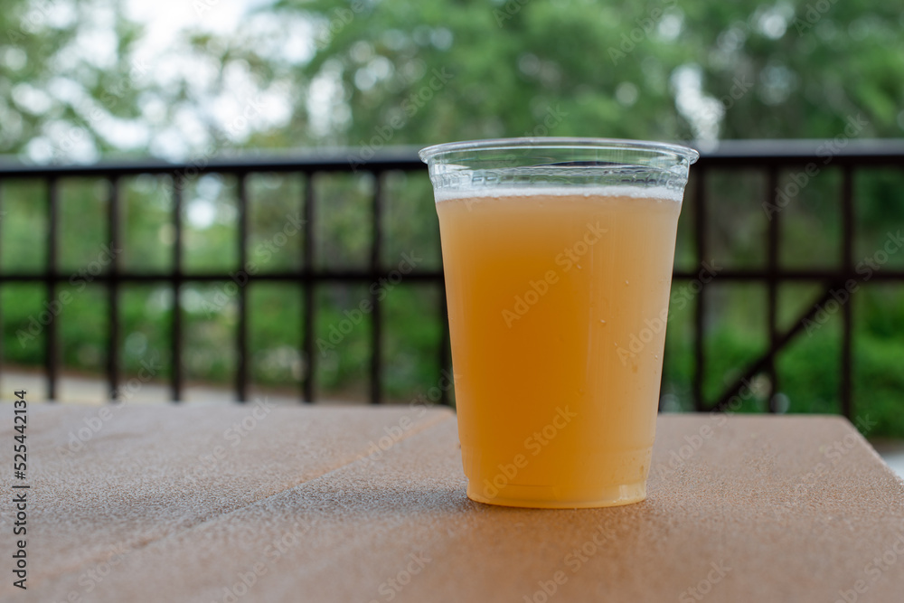 Tall clear plastic glass of sour craft beer in a microbrewery. The cold refreshment has frost on the glass. The beer is on a wooden patio table. The top of the glass has white froth around the rim. Tall clear plastic glass of sour craft beer in a microbrewery. The cold refreshment has frost on the glass. The beer is on a wooden patio table. The top of the glass has white froth around the rim.