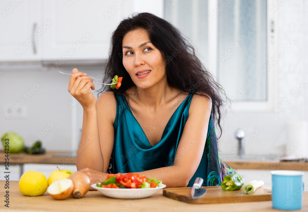 Portrait of positive asian woman in nightie eating salad in kitchen at home. Woman has cooked fresh vegetable salad.