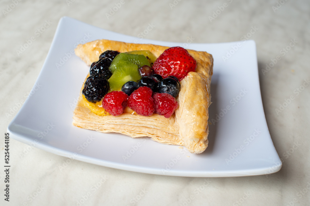 A square fruit filled French puff pastry on a white square plate. The cream and custard filled dessert have strawberries, kiwi, and blackberries on top with a sugar glaze. The pastry is flaky. A square fruit filled French puff pastry on a white square plate. The cream and custard filled dessert have strawberries, kiwi, and blackberries on top with a sugar glaze. The pastry is flaky.
