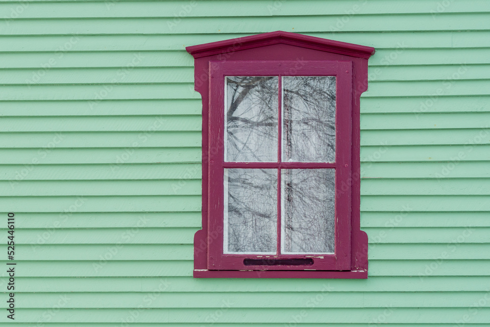 A single four pane Victorian style glass window with a deep pink decorative trim. There's a lace curtain on the inside. The window is in a vibrant mint green narrow clapboard exterior wall of a house. A single four pane Victorian style glass window with a deep pink decorative trim. There's a lace curtain on the inside. The window is in a vibrant mint green narrow clapboard exterior wall of a house.