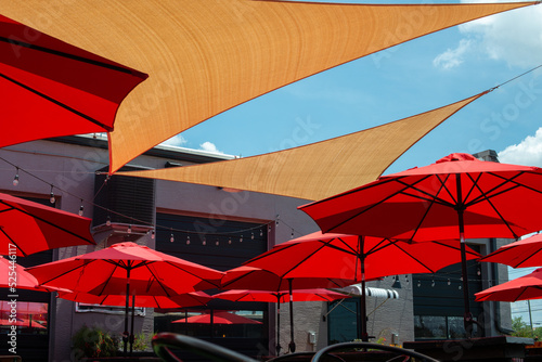 Multiple triangle shaped yellow nylon sunshades and awnings hanging over a patio deck. There are red colored canvas umbrellas hung with strings of clear patio light against a bright blue sunny sky. photo