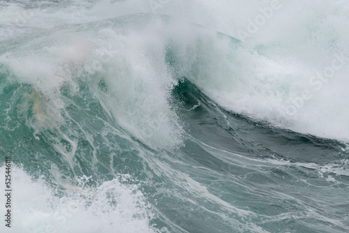 An angry turquoise green color massive rip curl of a wave as it barrels rolls along the ocean. The white mist and froth from the wave are foamy and fluffy. The ocean in the background is deep blue. 