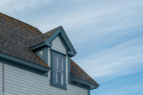 The corner of a vintage traditional wooden house with white clapboard and blue trim. There's a peaked dormer window on the roof. The roof is covered in cedar shakes with blue sky and clouds.
