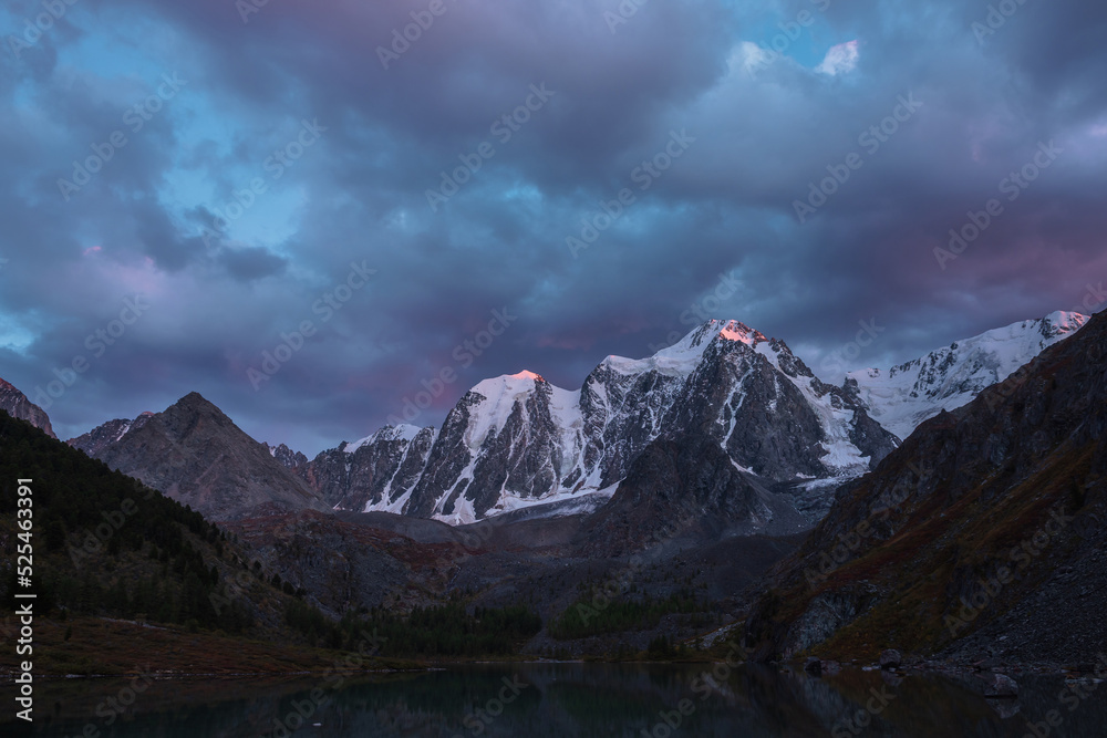 Obraz premium Atmospheric autumn twilight landscape with sunset gold reflection on huge snowy mountain top in violet dramatic sky. Mountain lake with view to giant snow-covered mountains under cloudy sky in dusk.