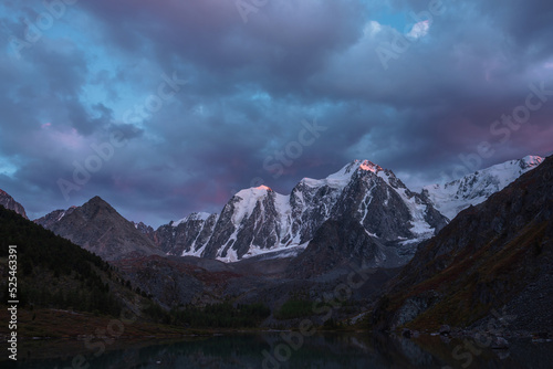 Atmospheric autumn twilight landscape with sunset gold reflection on huge snowy mountain top in violet dramatic sky. Mountain lake with view to giant snow-covered mountains under cloudy sky in dusk.