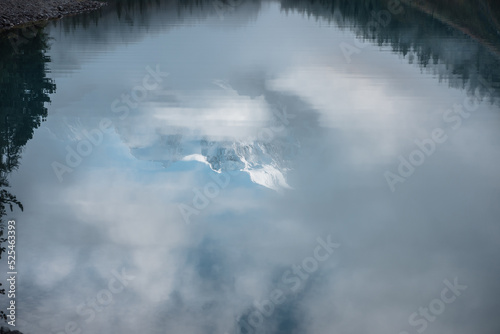 Tranquil scenery with reflex in mountain lake of snow castle in clouds. Snowy mountains in fog clearance and coniferous trees reflected in alpine lake. Beautiful landscape with mirror of glacial lake.