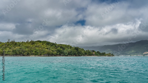 The tropical island is completely overgrown with green vegetation. Turquoise ocean and white cumulus clouds in the sky. Seychelles