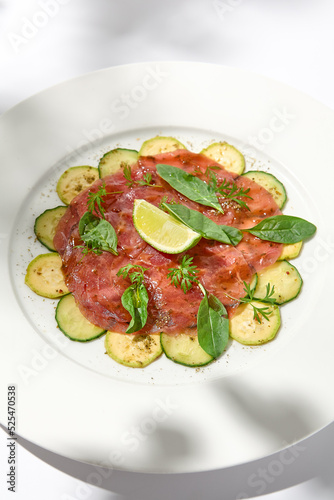 Beef carpaccio with zucchini and cucumber on white table with sunlight shadows. Appetizer of meat - beef carpaccio on summer dining. Sliced raw meat in italian style. Aesthetic menu.