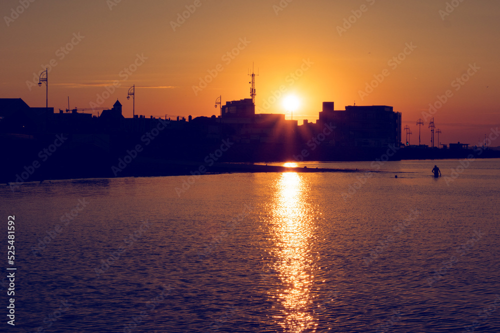 Silhouette of a town building at sunrise. Warm orange color. Calm and peaceful mood. Start of a new day concept. Galway city, Ireland. Sun glow in the ocean surface.