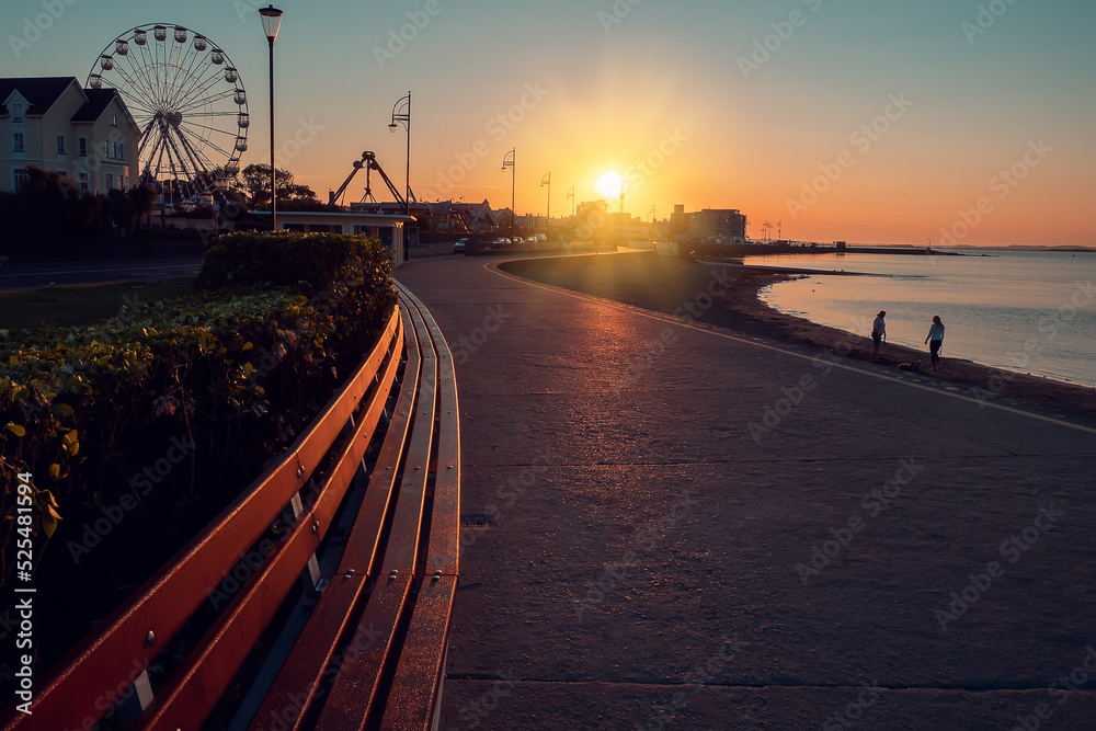 Sunrise scene at Salthill promenade in Galway city, Ireland. Long wooden sitting bench. Town buildings and fun fair wheel silhouette. Sun flare. Warm orange colors. Calm mood.