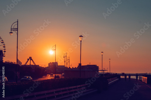 Sunrise scene at Salthill promenade in Galway city, Ireland. Town buildings and fun fair wheel silhouette, Sun in place of town light. Warm orange colors. Calm mood.