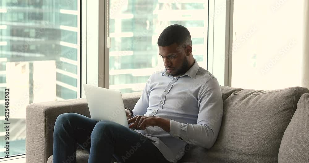 Handsome African man sit on sofa, using modern wireless computer, working in modern skyscraper flat, typing e-mail on laptop keyboard looks focused. Workflow, lifestyle, business, technology concept Handsome African man sit on sofa, using modern wireless computer, working in modern skyscraper flat, typing e-mail on laptop keyboard looks focused. Workflow, lifestyle, business, technology concept