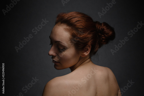 Pretty tan woman with ginger hair close her eyes and posing on a camera. The view from the back. Dark background