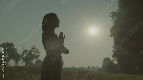 Silhouette of elegant attractive African American woman in sundress holding hands in prayer, praying for god bless and mercy while standing on green field over beautiful summer sunrise. photo