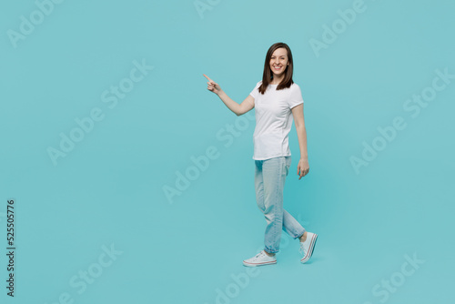 Full body side view young caucasian woman 20s wear white t-shirt walking going strolling point index finger aside on workspace area mock up isolated on plain pastel light blue cyan background studio.