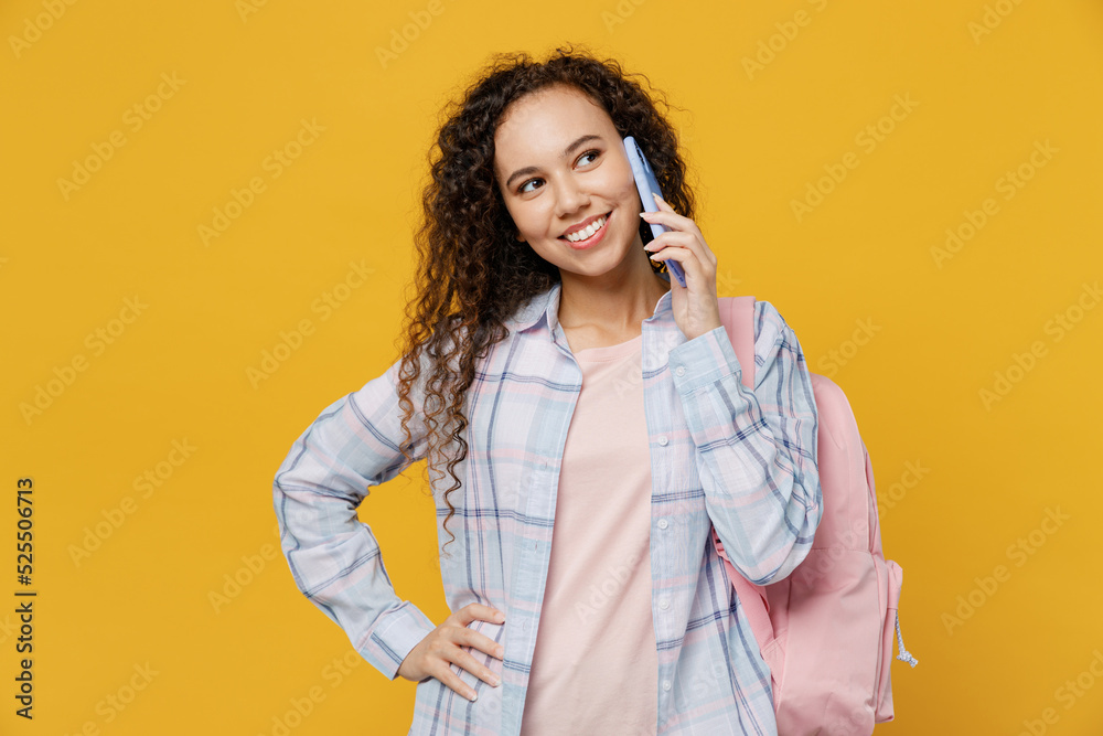 Young smiling happy fun black teen girl student she wear casual clothes backpack bag talk speak on mobile cell phone isolated on plain yellow color background. High school university college concept. Young smiling happy fun black teen girl student she wear casual clothes backpack bag talk speak on mobile cell phone isolated on plain yellow color background. High school university college concept.