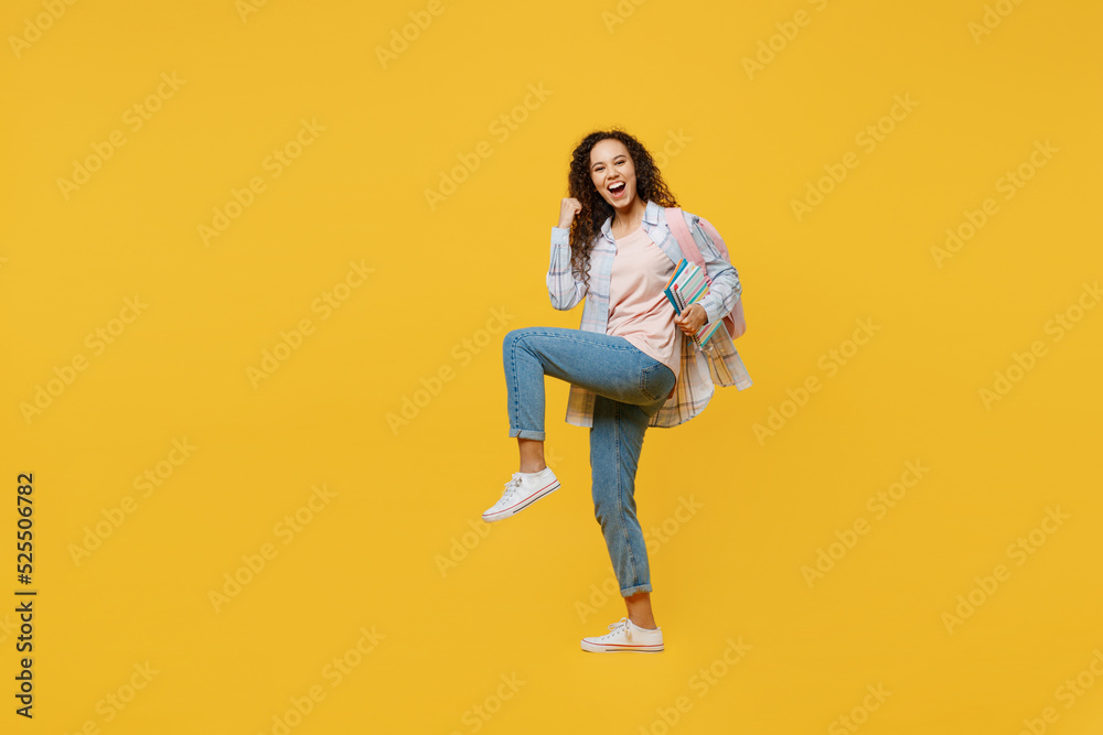Full body young black teen girl student she wear casual clothes backpack bag hold books do winner gesture clench fist isolated on plain yellow color background. High school university college concept.