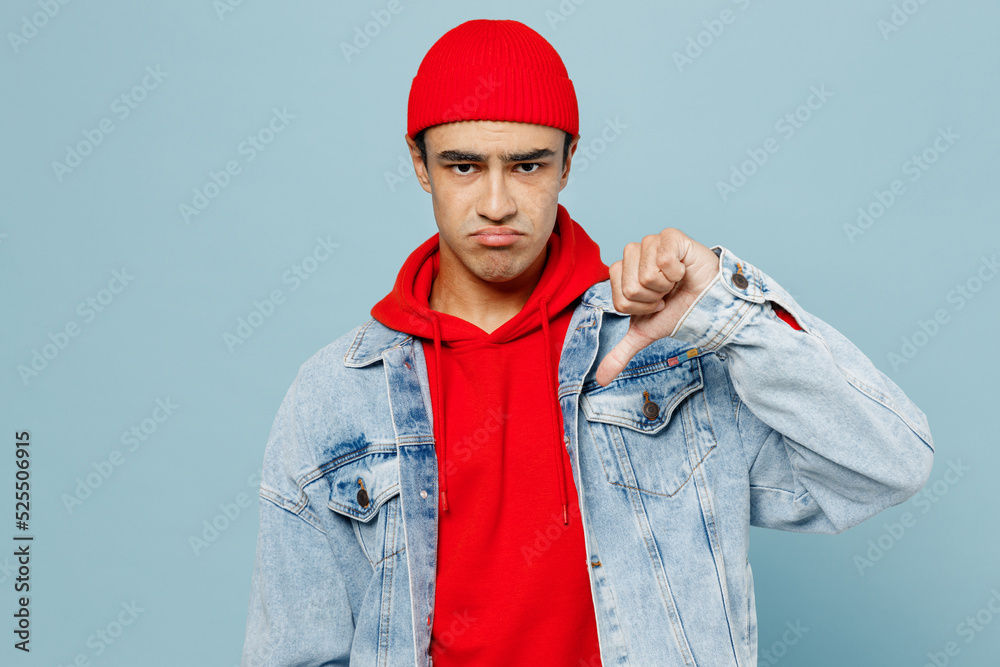 Fototapeta premium Young sad unhappy displeased middle eastern man 20s he wear denim jacket red hat showing thumb down dislike gesture isolated on plain pastel light blue cyan background studio People lifestyle concept